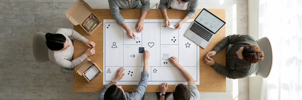 Großer Workshop zur Markenstrategie: Mehrere Personen sitzen und stehen um einen Tisch, betrachten Whiteboard mit Kreisen und Pfeilen, Laptop mit Shop-Skizze und Karten mit Markenwerten.