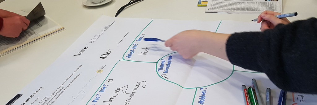 Hand füllt in einem Design Thinking Workshop ein großes Persona-Poster mit Feldern wie Name, Alter, Arbeit und Träume aus, daneben liegen Stifte auf dem Tisch.