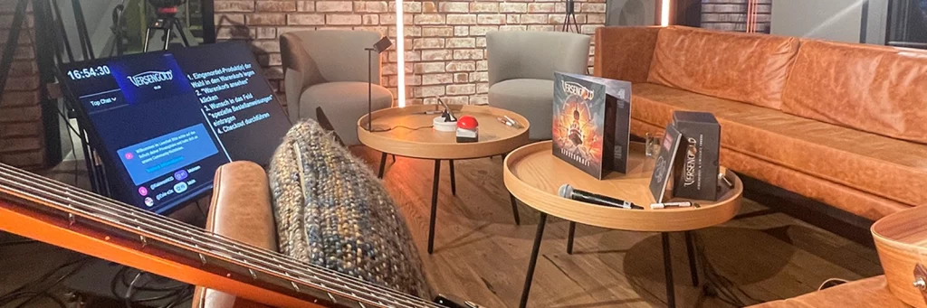 Detailaufnahme des YouTube-Live-Sets: Couch, Sessel, runde Tische mit Album-Boxen und Mikrofon, im Hintergrund ein Screen mit Stream-Vorschau – vorbereitetes Markenerlebnis im Studio.