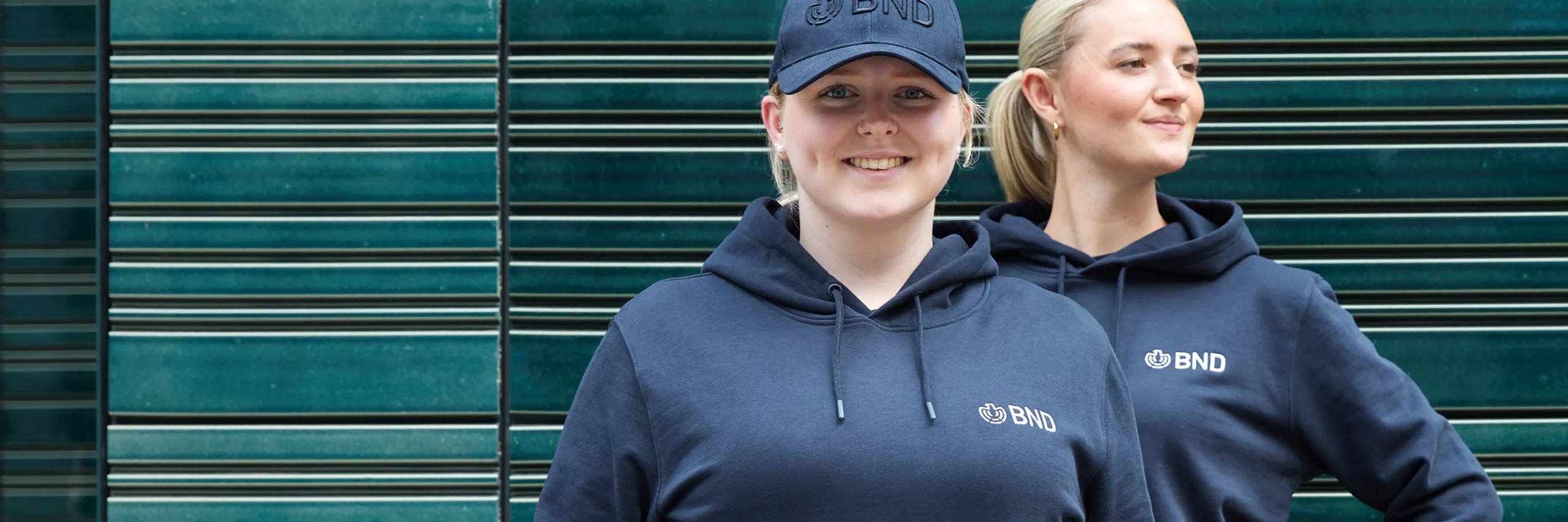 Zwei Frauen tragen dunkelblaue Hoodies aus dem BND Online Shop vor einer modernen, grünen Gebäudefassade.