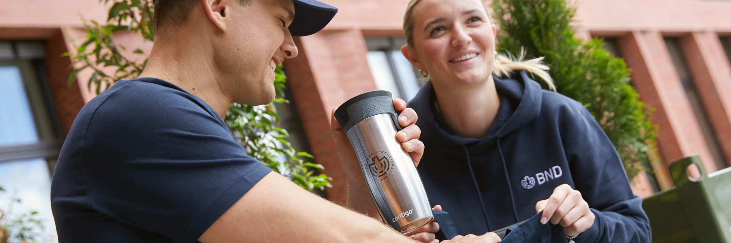Zwei Personen unterhalten sich entspannt, während eine Person einen Thermobecher aus dem BND Online Shop in der Hand hält.